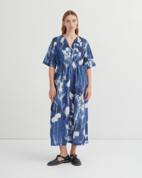 A woman stands against a plain light gray studio background wearing a loose, short-sleeve midi dress with a blue and white floral cyanotype print. The dress has a V-neck and gathered waist.
