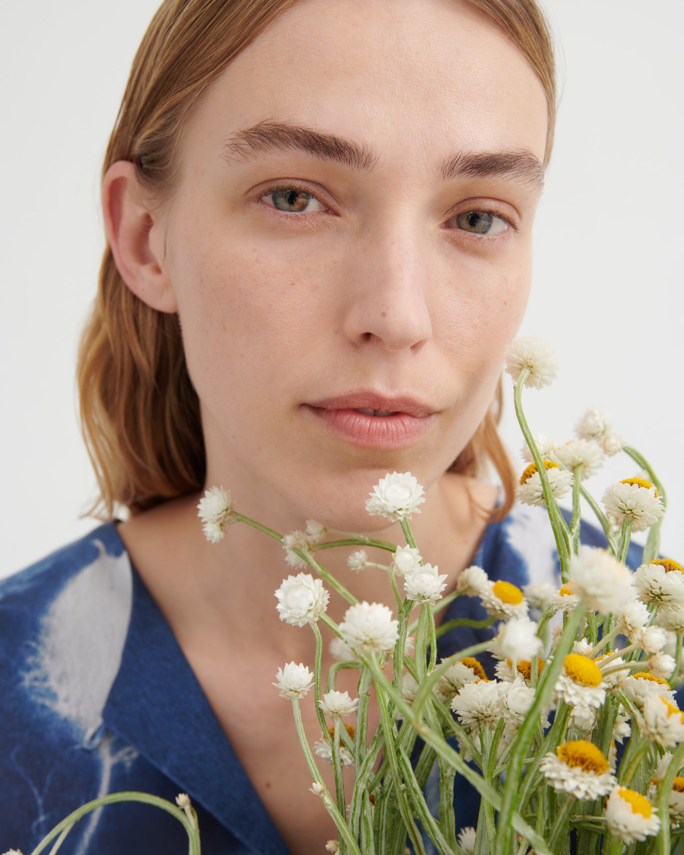 A close-up portrait of a woman with light brown hair wearing a blue shibori-patterned top, holding a bouquet of white and yellow wildflowers against a plain white background.