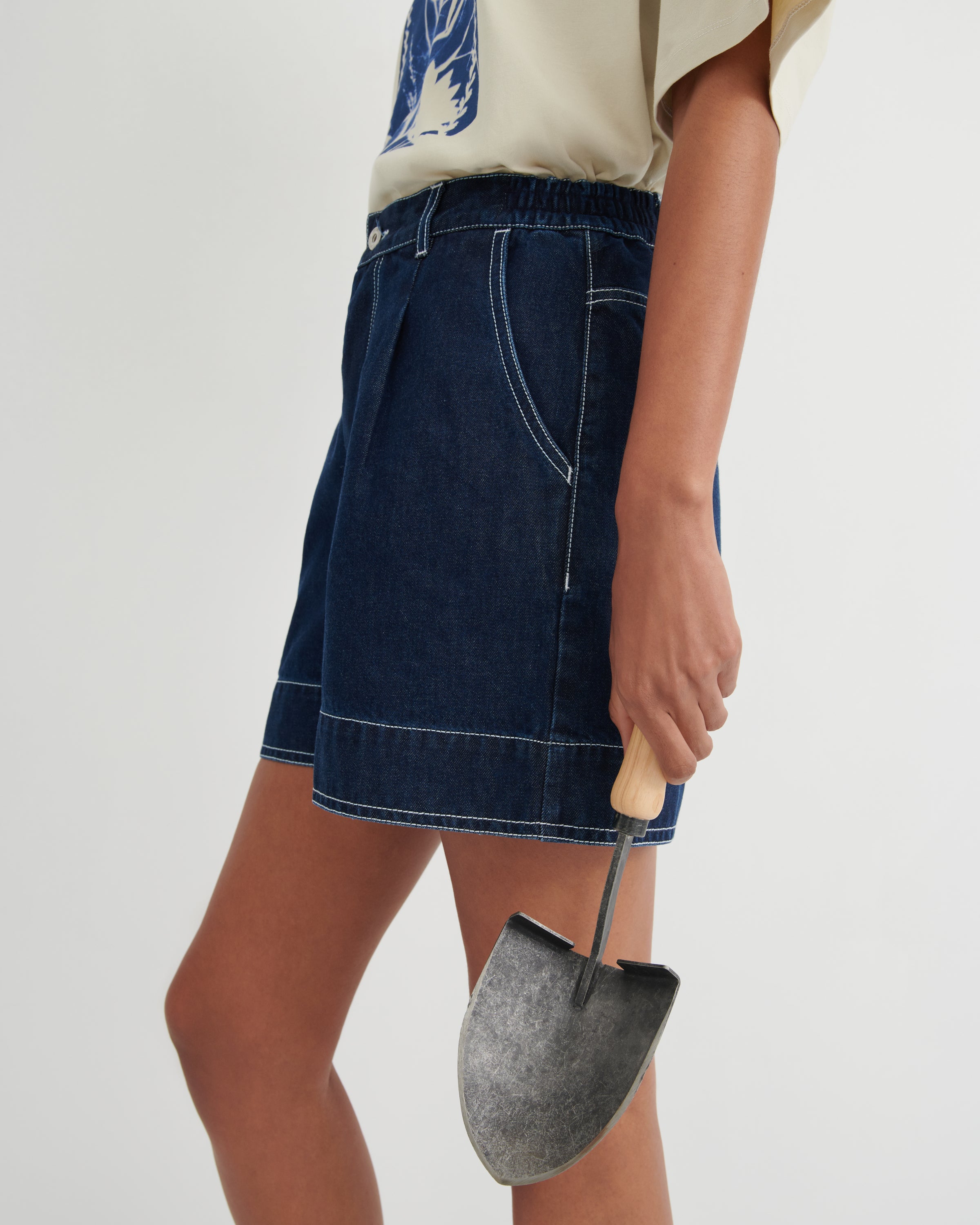 A model wearing a dark blue denim mini skirt with contrast white stitching, holding a small metal trowel, against a plain light gray studio background.