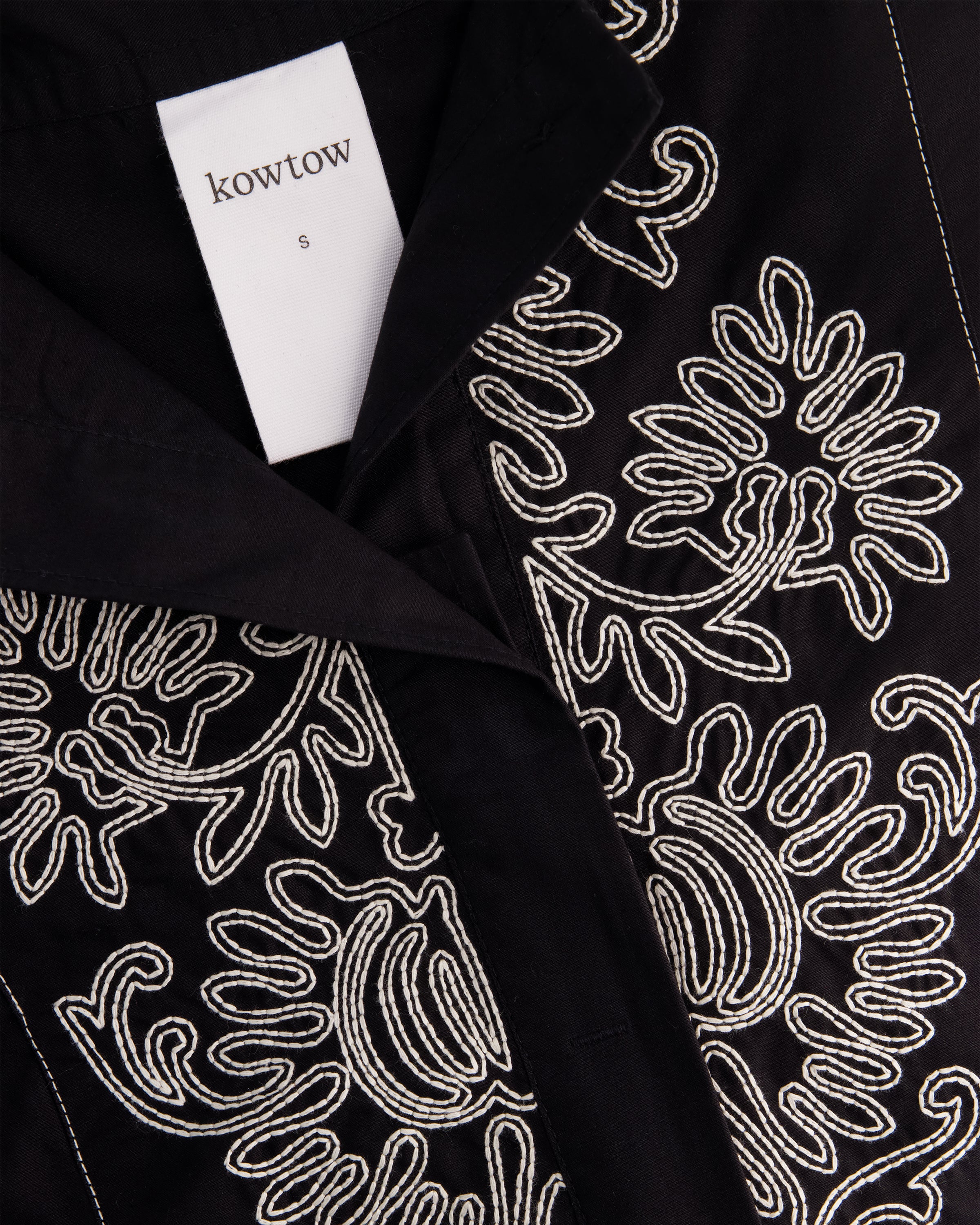 Close-up of a black garment with intricate white embroidered floral and paisley patterns, featuring a white brand label that reads 'kowtow'.