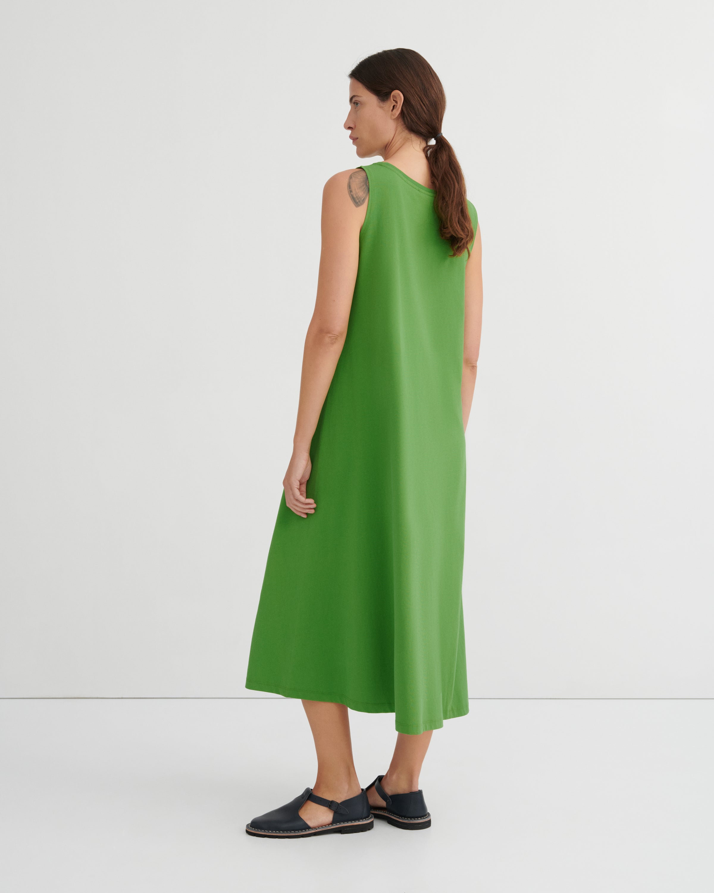 A woman wearing a sleeveless bright green midi dress, shown from the back in a studio setting with a white background.