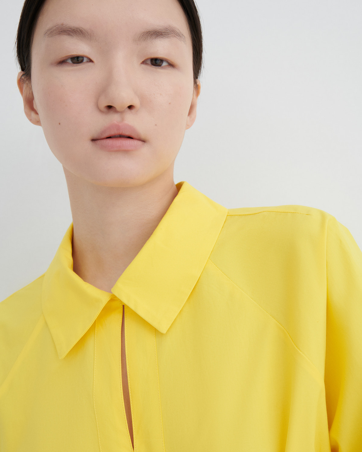 A close-up of a model wearing a bright yellow collared blouse against a plain light gray background.