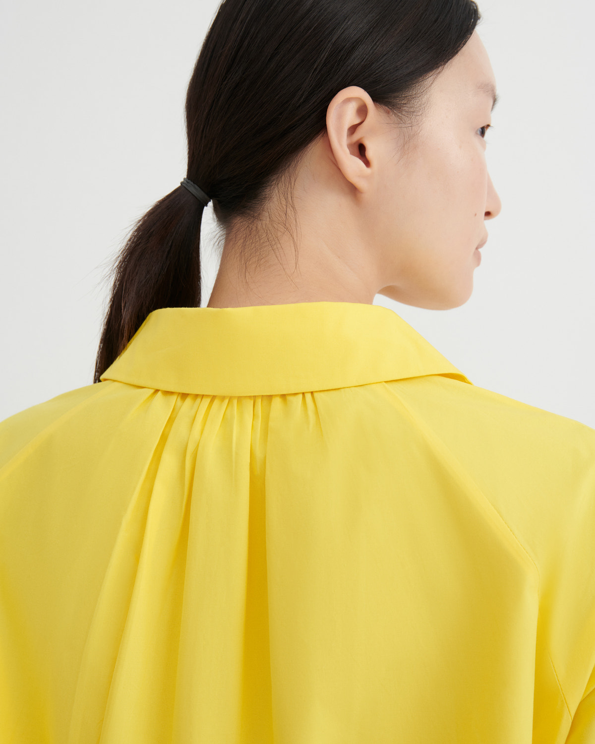 Close-up side profile of a model wearing a bright yellow blouse with a wide collar and gathered neckline, against a plain light gray background.
