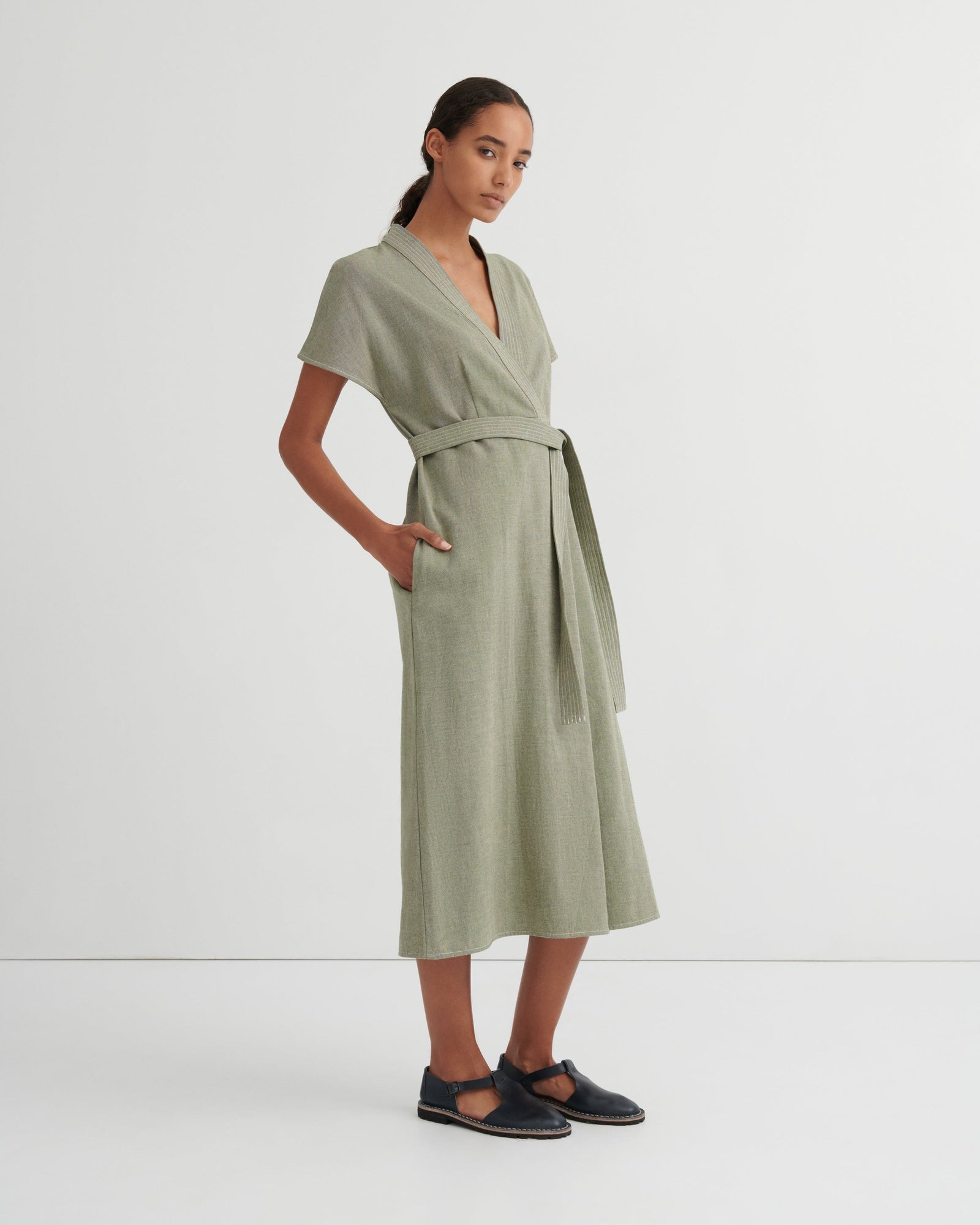 A woman modeling a sage green linen wrap dress with short sleeves, a V-neckline, a matching waist tie, and side pockets, paired with dark navy Mary Jane flats, against a plain white studio background.