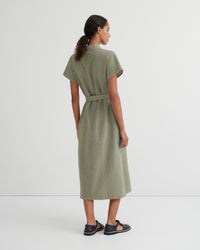 A woman shown from the back wearing a short-sleeved olive green midi dress with a matching waist tie, paired with dark sandals, against a plain light gray studio background.