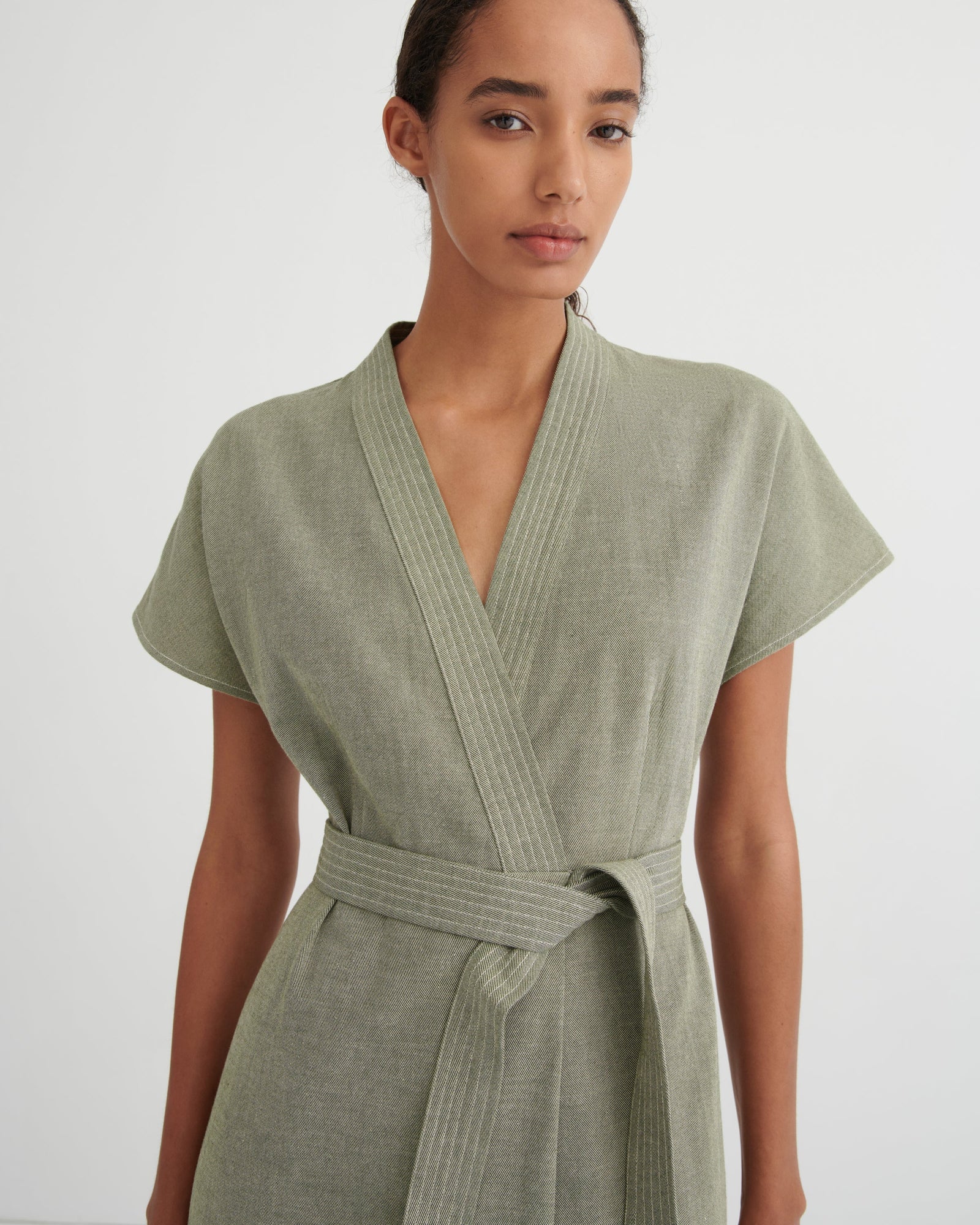 A woman wearing a sage green short-sleeve wrap dress with a matching tie belt, photographed against a plain light gray studio background.