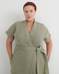 A woman wearing a sage green short-sleeve wrap robe with a matching waist tie, standing against a plain light gray background.