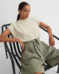 A woman seated on a black metal chair wearing a cream short-sleeve mock-neck top and olive green high-waisted wide-leg shorts with a tie belt, against a clean white studio background.