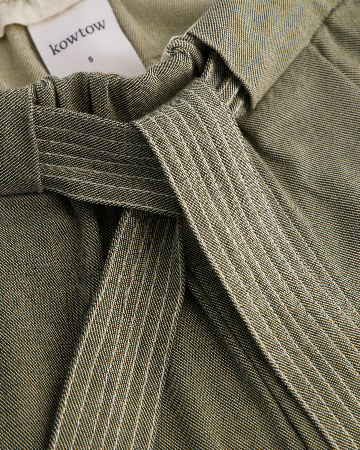 Close-up of a Kowtow garment in olive green fabric with a matching tie belt and a white brand label visible at the collar.