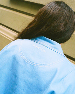 A person viewed from behind wearing a light blue Kowtow overshirt, with their dark brown hair visible against a beige wall background.