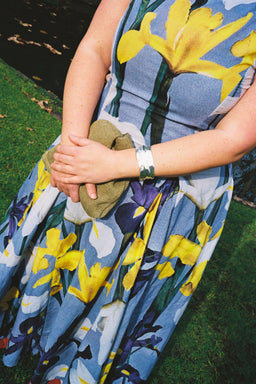 A close-up of a person outdoors wearing a sleeveless blue dress with a large yellow, purple, and white floral print. The person is holding a small olive-green clutch and wearing a silver cuff bracelet, set against a background of green grass.