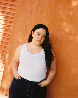 A woman with long dark hair stands against a textured orange wall, wearing a white tank top and dark blue jeans.