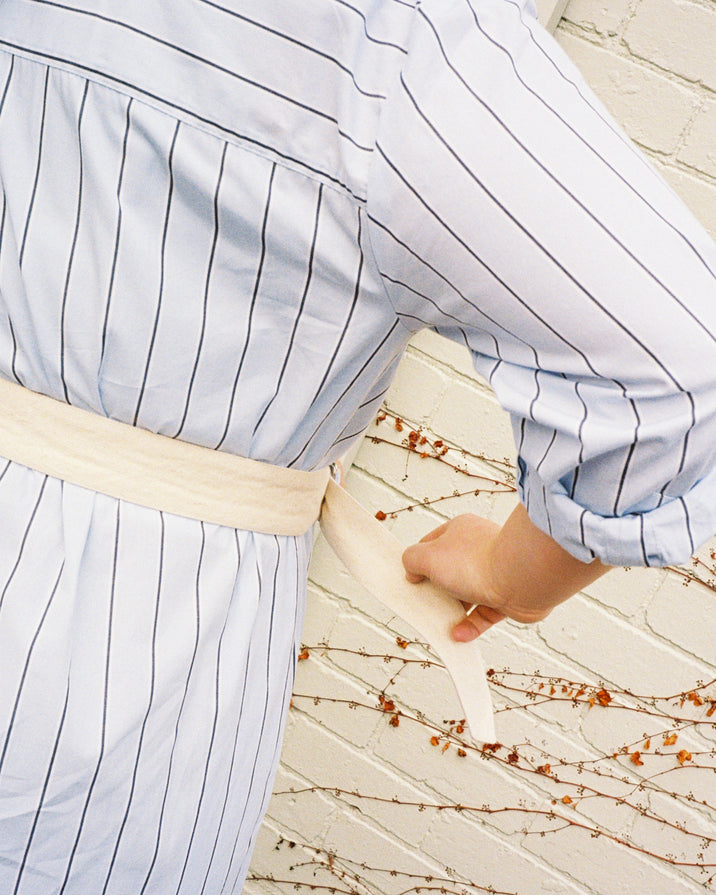 A close-up lifestyle shot of a person wearing a light blue and white pinstriped shirt with a cream-colored waist belt.