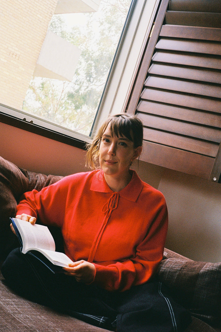 A woman with bangs sits on a couch by a window, wearing a bright orange-red long-sleeve collared top and reading an open book. Wooden shutters are visible next to the window.