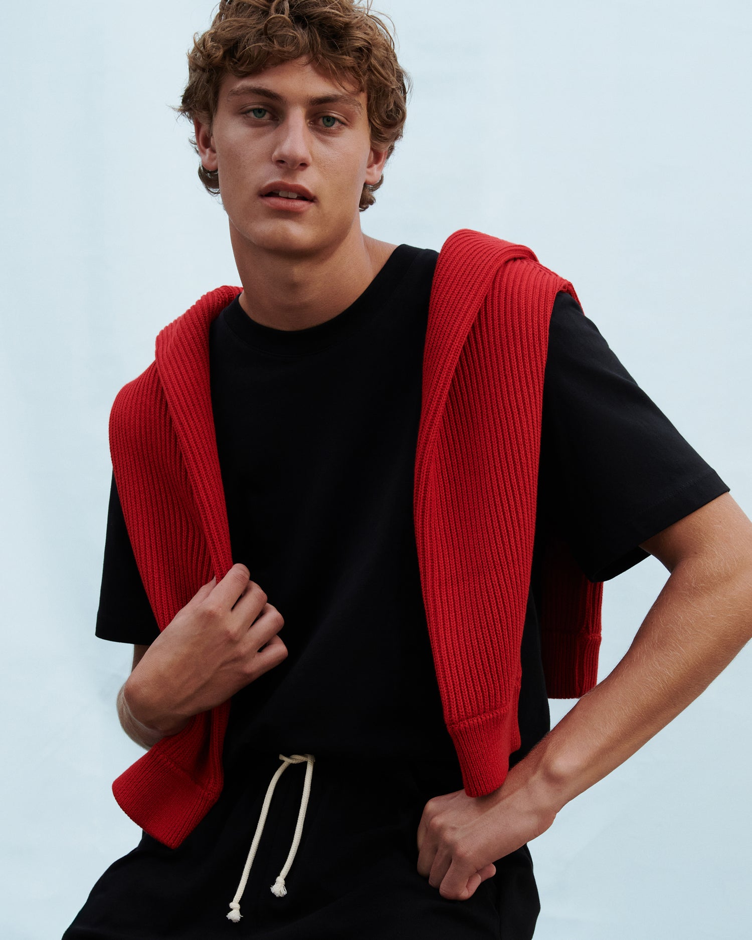 Model against a blue background, wearing a relaxed black t-shirt and a red sweater draped over his shoulders.