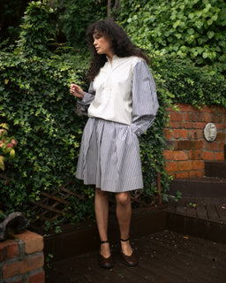 A woman stands outdoors on a wooden deck in front of a brick wall and dense green ivy, wearing a cream button-front vest over a blue-and-white vertically striped long-sleeve blouse and matching striped pleated skirt, with dark brown ankle-strap shoes.