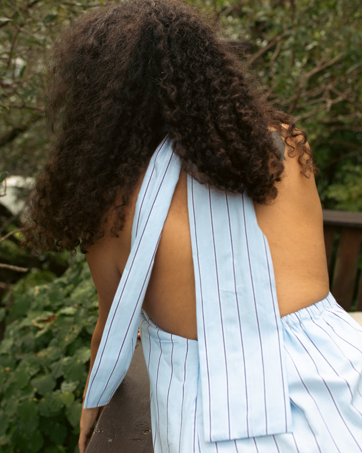 A person outdoors wearing a light blue and white striped halter dress, shown from the back with curly dark hair and a cutout back design.