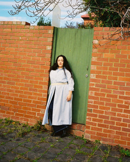 A woman stands in front of a red brick wall and a green wooden door, wearing a long white and blue striped dress with a beige belt. Bare tree branches hang from above, and the ground is paved with cobblestones.