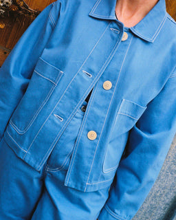 A close-up of a person wearing a bright blue work jacket with white contrast stitching and light-colored buttons, paired with matching blue pants.