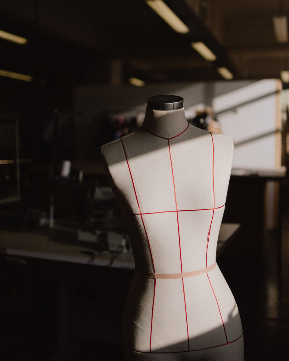 A cream-colored dress form mannequin with red measurement guide lines, standing in a dimly lit sewing studio with dramatic side lighting.