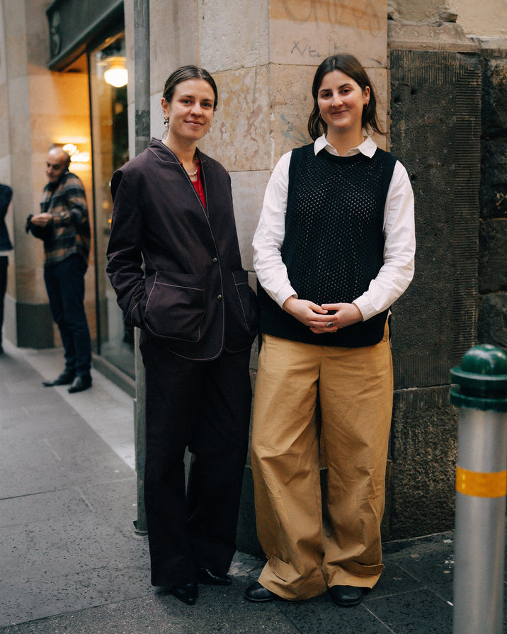 Two women standing on a city sidewalk in front of a stone building. The woman on the left wears a dark blazer and trousers over a red top, and the woman on the right wears a black knit vest over a white shirt with wide tan pants.