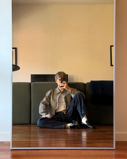 A man sits cross-legged on a wooden floor in front of a green sofa, taking a mirror selfie. He is wearing a beige and brown plaid button-down shirt over a light beige undershirt, dark navy trousers, white socks, and black shoes.