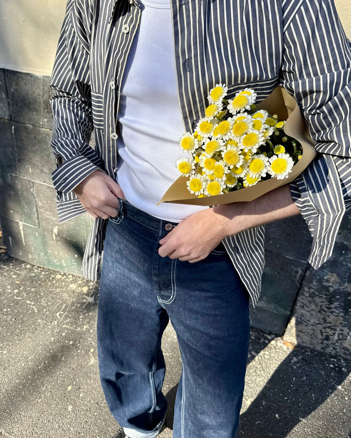 A person outdoors wearing a black-and-white vertically striped button-up shirt over a white t-shirt, blue jeans, and black shoes, while holding a bouquet of yellow and white daisies wrapped in brown paper.