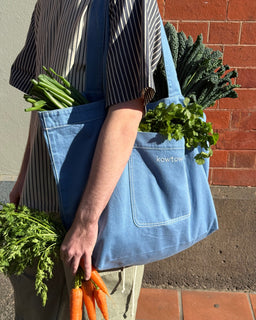 A person wearing a striped shirt carries a large light blue canvas tote bag filled with greens and produce, standing outdoors against a red brick wall. The bag has a front pocket with the brand name 'KOWTOW' printed in white.