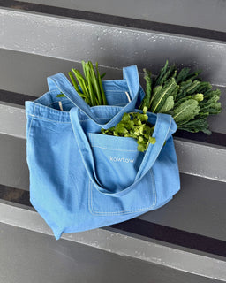 A light blue canvas tote bag with the brand name 'kowtow' printed on the front pocket, filled with green vegetables, resting on gray wooden slats.