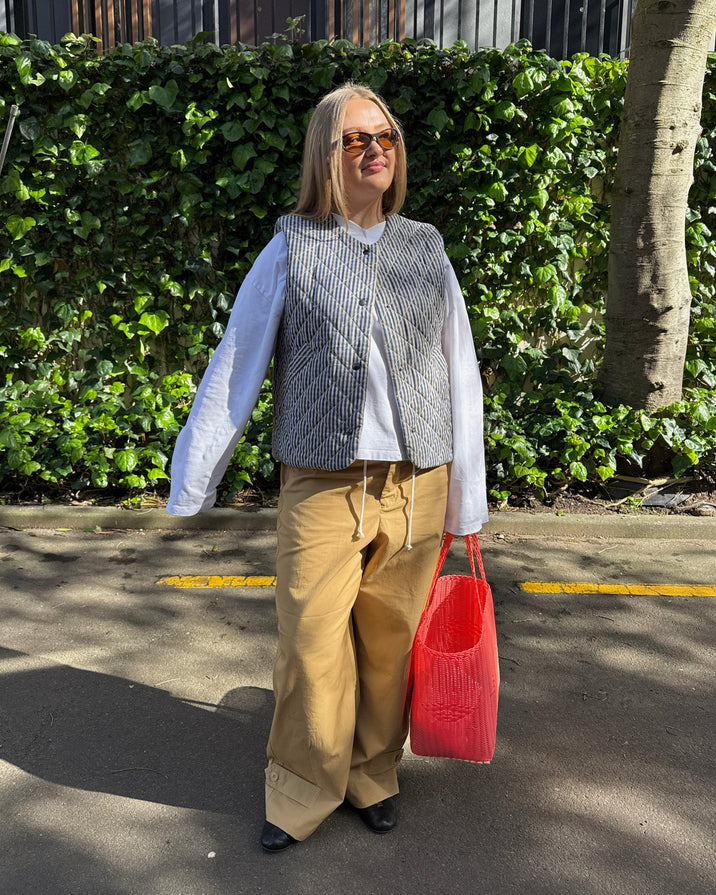 A woman stands outdoors on a paved path in front of a green hedge, wearing a gray quilted vest over a white long-sleeve shirt, tan wide-leg pants, and holding a bright pink woven tote bag.