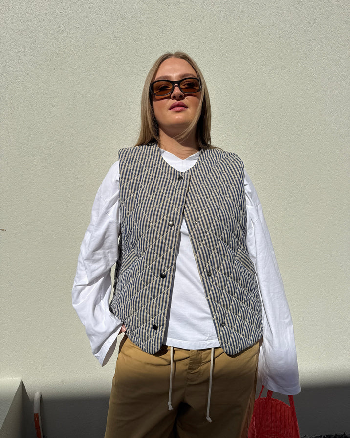 A woman wearing a gray and white striped quilted vest over a white long-sleeve shirt, tan pants, and dark sunglasses, standing against a plain light beige wall.