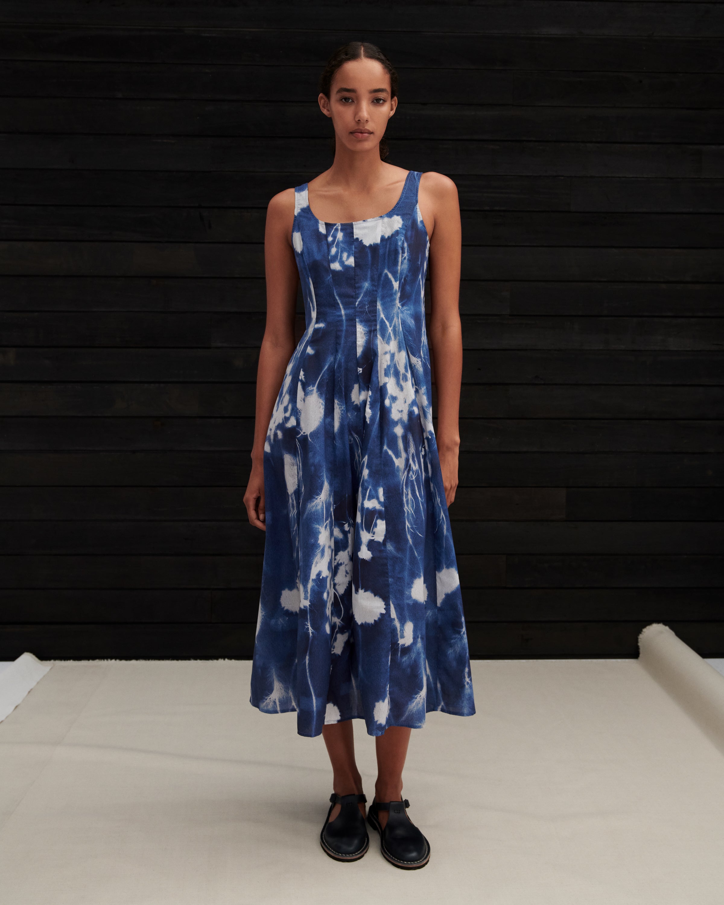 A woman modeling a sleeveless midi dress with a blue and white tie-dye pattern, standing against a dark wood-paneled wall.