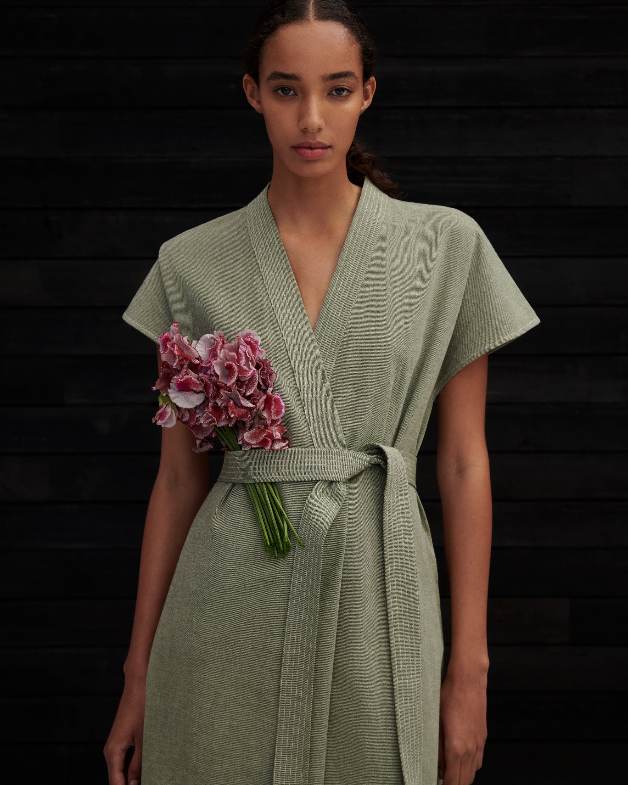 A woman wearing a sage green linen wrap robe with a matching tie belt, holding a bouquet of pink flowers against a dark wood plank background.