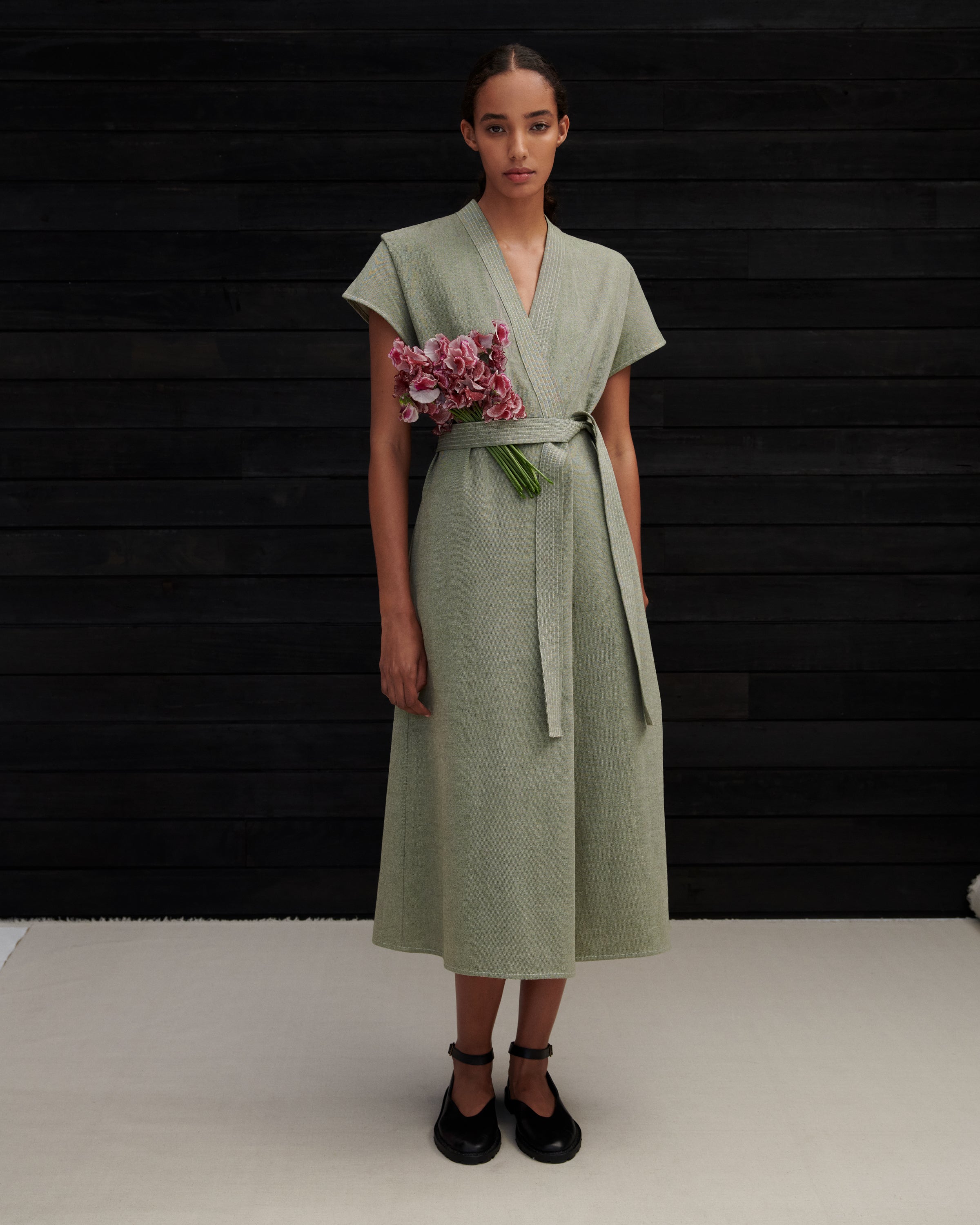 A model stands against a dark wood-paneled wall wearing a sage green wrap dress with a matching waist tie belt and black Mary Jane shoes, holding a small bouquet of pink flowers.