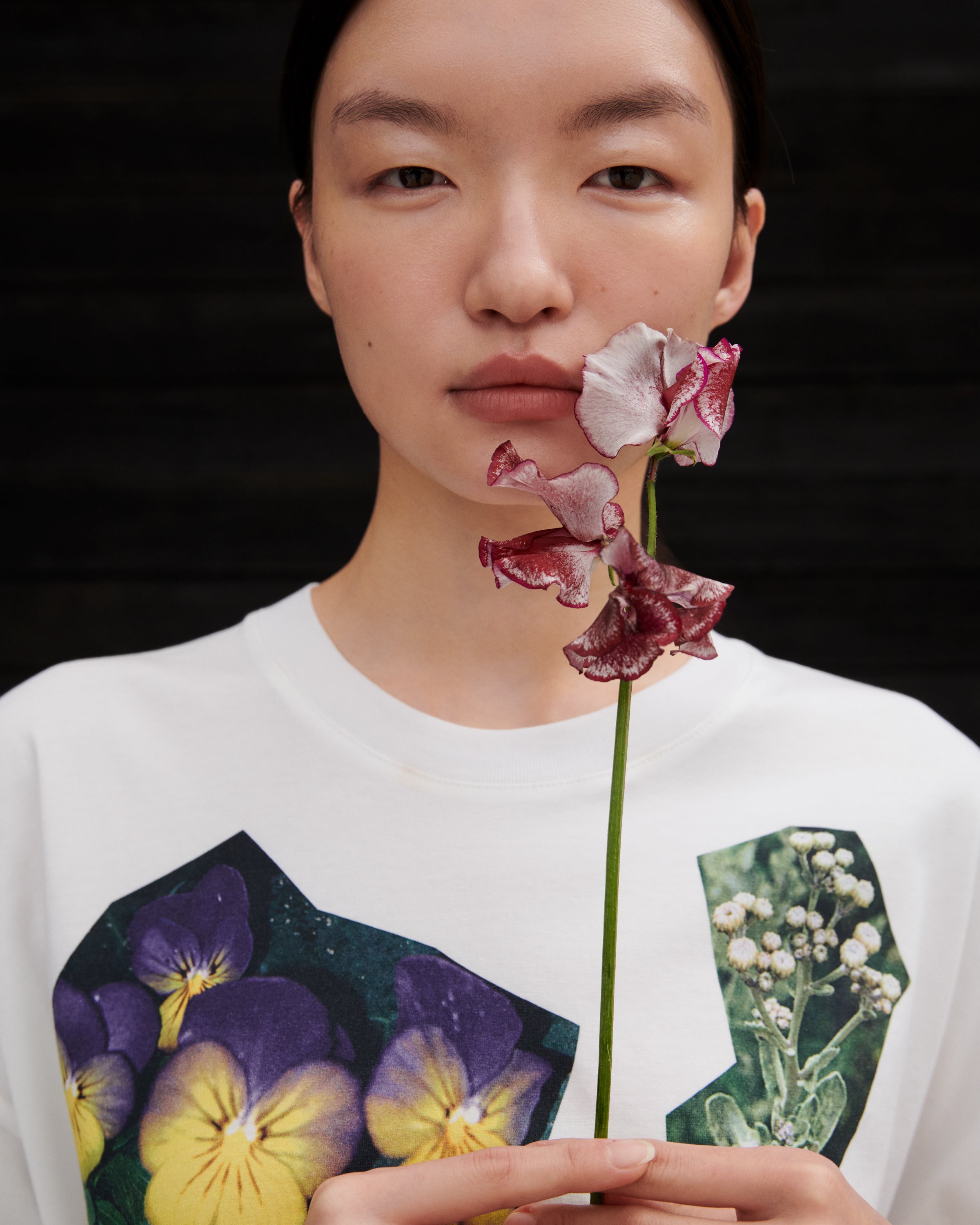 A model wearing a white crewneck sweatshirt with a graphic print of purple pansies and baby's breath, holding a flower stem in front of their face against a dark background.