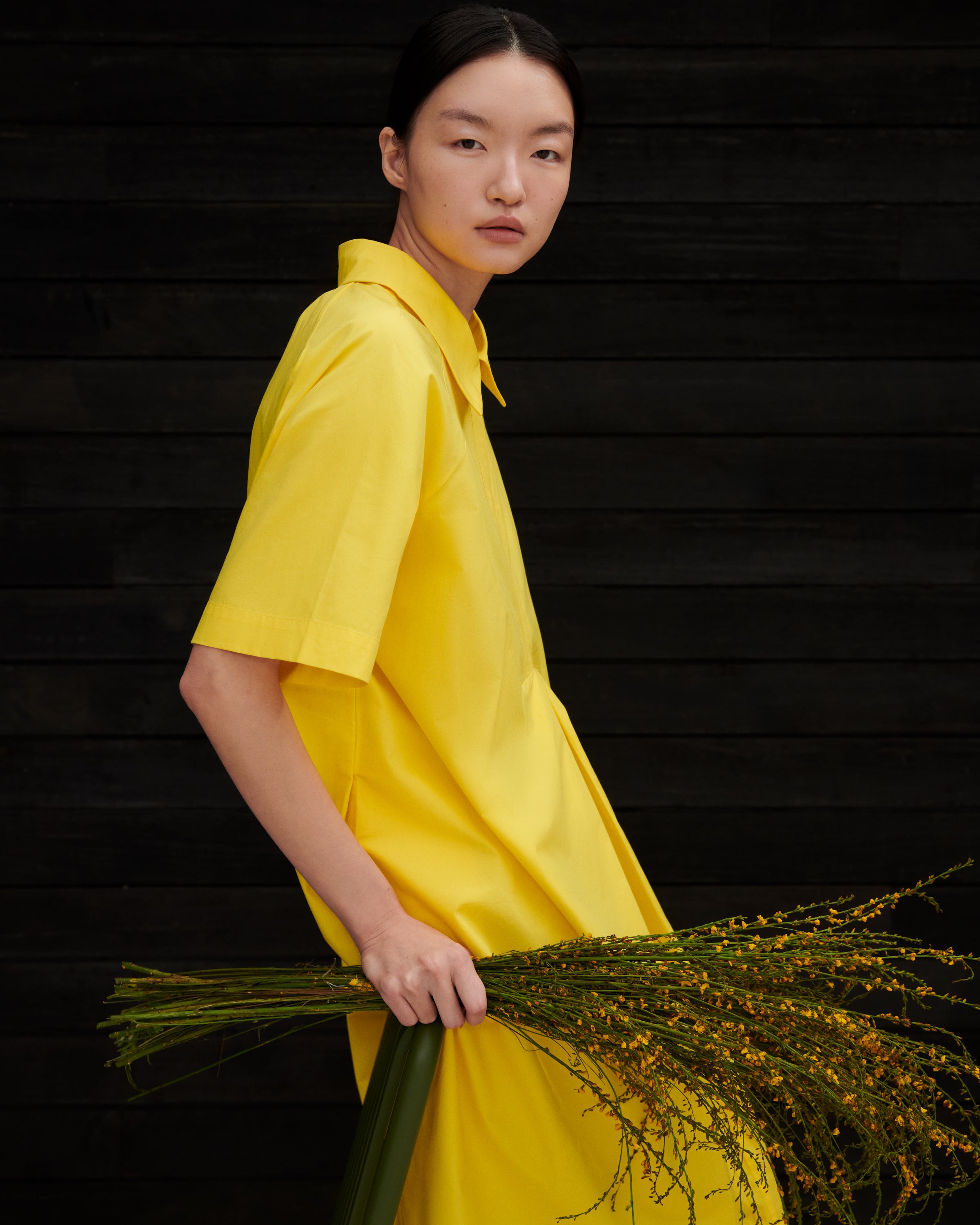 A model wearing a bright yellow short-sleeve collared shirt dress, holding a bunch of green and yellow grasses against a dark wood plank background.