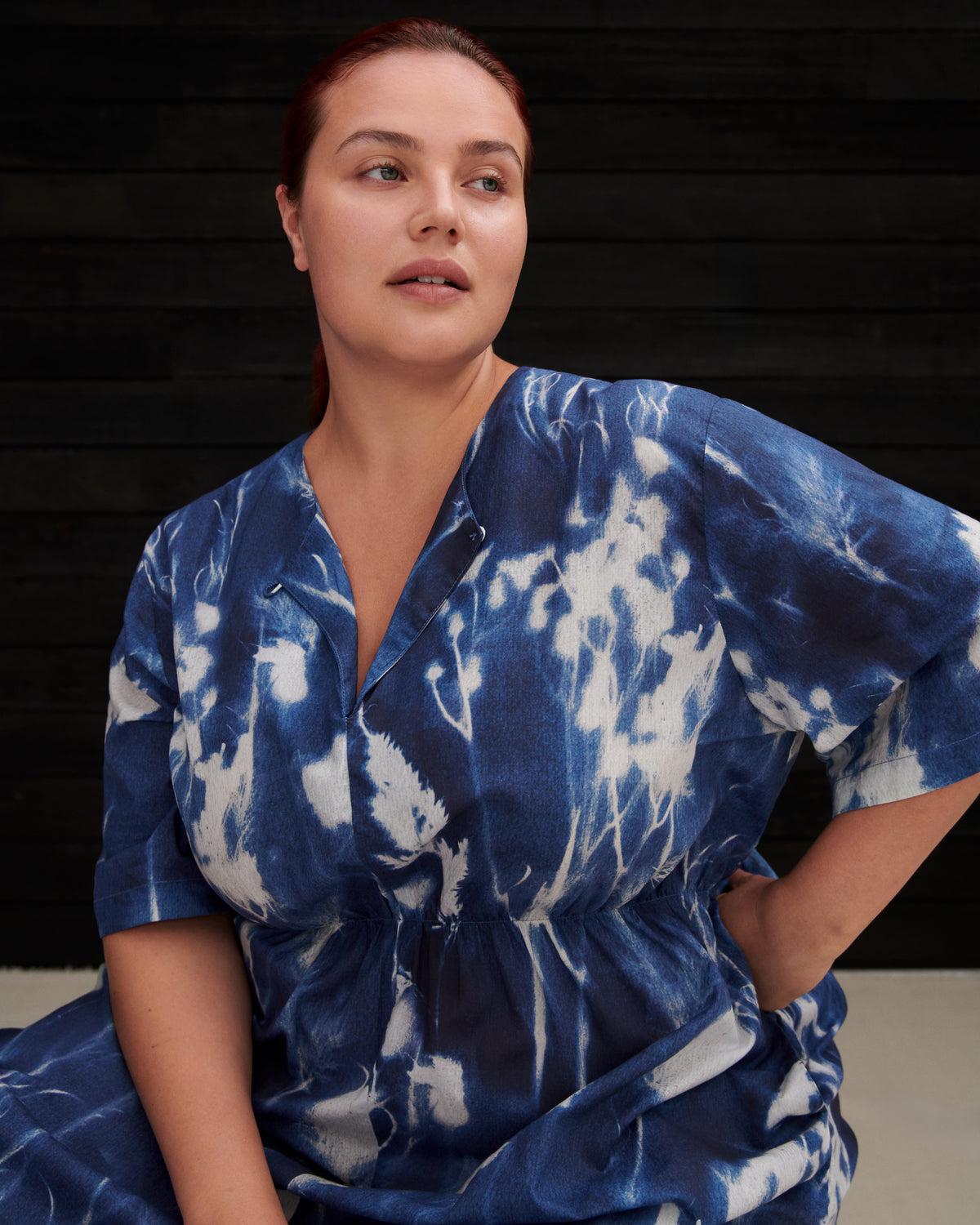 A woman wearing a blue and white tie-dye short-sleeve wrap dress, posed against a dark wooden background.