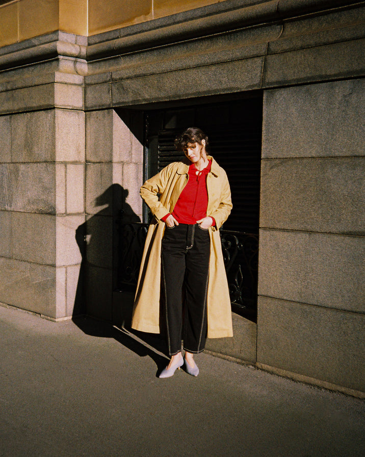 A person stands in a stone building doorway wearing a long beige trench coat over a bright red top and black trousers, with strong sunlight casting a sharp shadow on the wall.