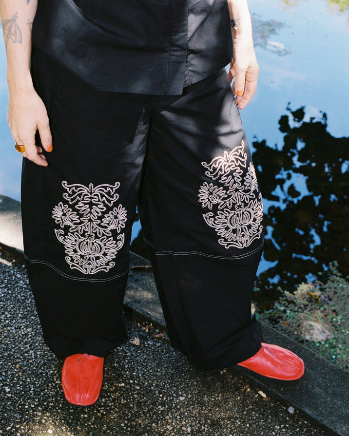 A person wearing black wide-leg pants with large white floral embroidery on the lower legs, paired with bright red shoes, standing outdoors near water.