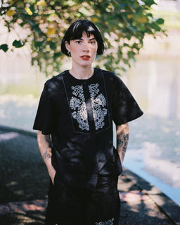 A person with short dark hair stands outdoors under leafy trees near a body of water, wearing a loose black dress with white floral embroidery around the neckline and on the lower front.