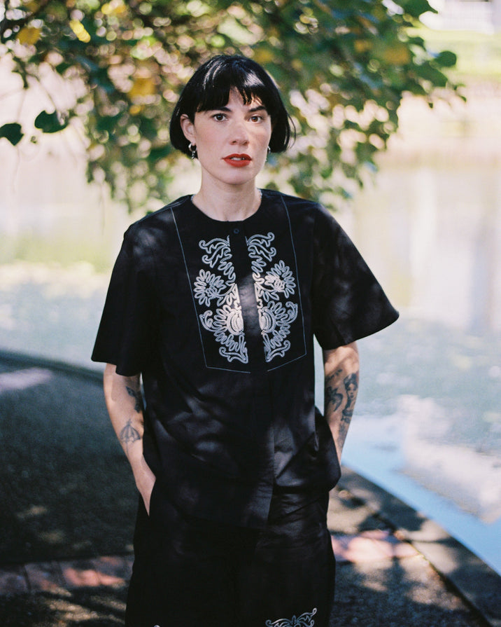 A person with short dark hair stands outdoors under leafy trees near a body of water, wearing a loose black dress with white floral embroidery around the neckline and on the lower front.