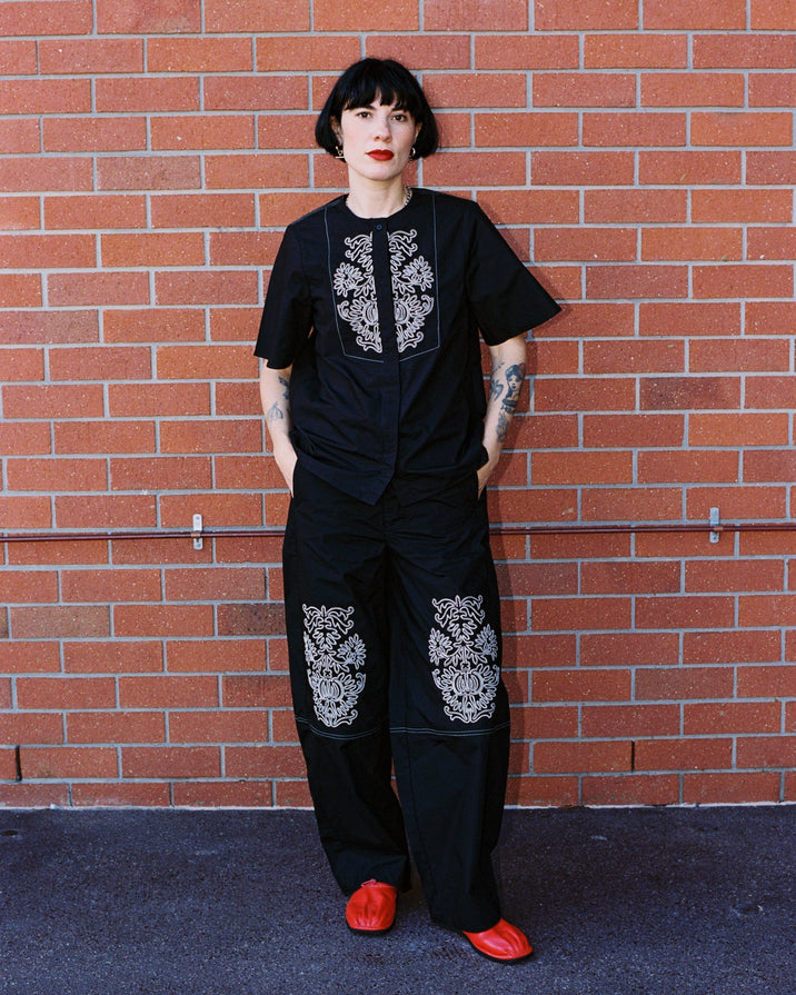 A person with short black hair and red lipstick stands against a red brick wall wearing a black short-sleeve top and matching wide-leg pants with white floral embroidery, paired with bright red shoes.