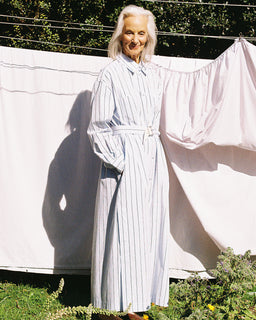 An older woman with white hair stands outdoors in front of hanging white sheets, wearing a long white shirt dress with thin blue vertical pinstripes and a matching belt. The scene is lit by bright natural sunlight, casting a strong shadow on the sheet.