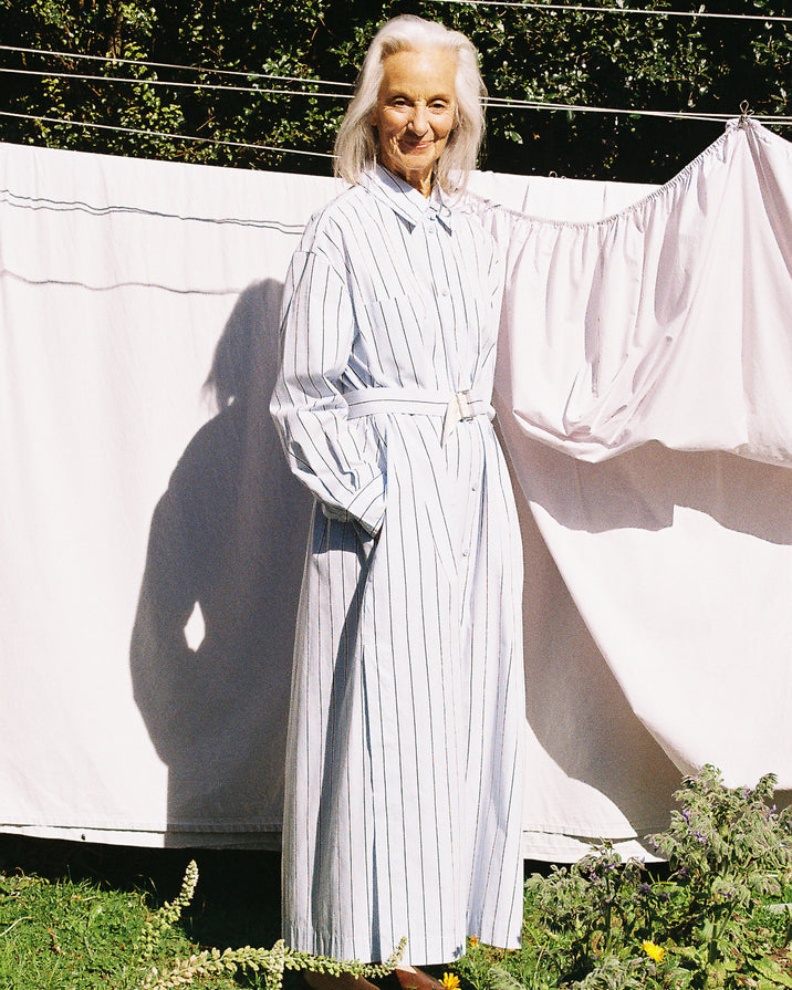 An older woman with white hair stands outdoors in front of hanging white sheets, wearing a long white shirt dress with thin blue vertical pinstripes and a matching belt. The scene is lit by bright natural sunlight, casting a strong shadow on the sheet.