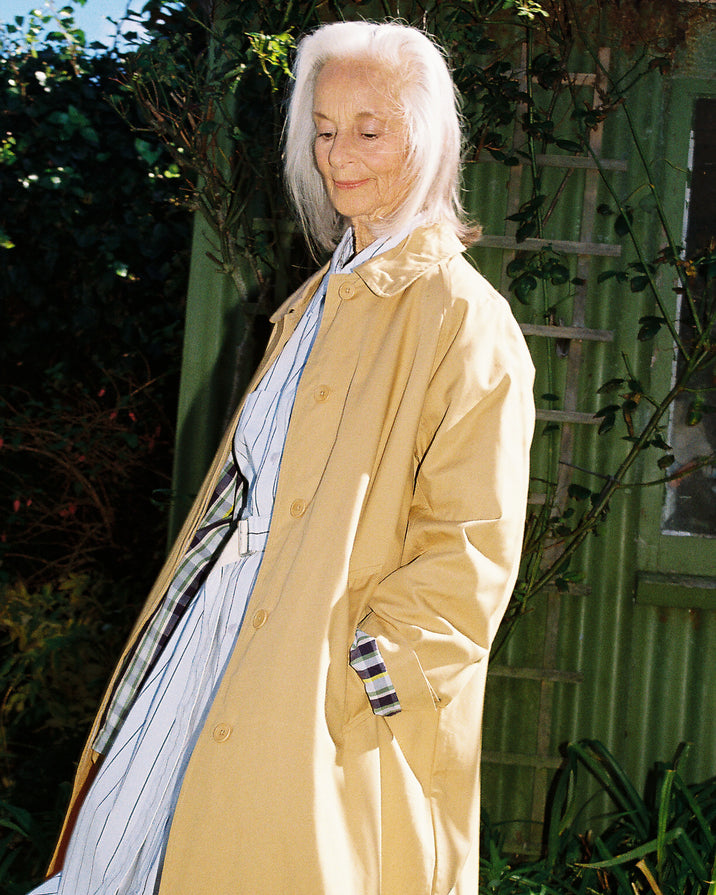 An elderly woman with white hair stands outdoors wearing a long beige trench coat over a blue and white striped dress, with a plaid pocket square in her hand, set against a green garden background.