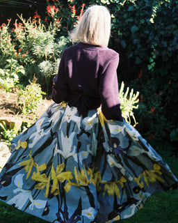 A woman viewed from behind in a sunlit garden, wearing a dark purple cardigan and a voluminous skirt with a blue, white, yellow, and black floral print.