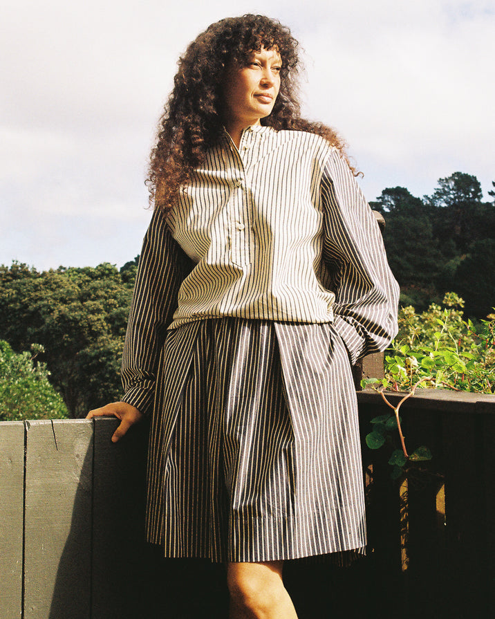 A woman with long curly hair stands outdoors wearing a matching striped button-front blouse and pleated skirt set. The outfit features vertical black and off-white stripes, set against a background of trees and a cloudy sky.