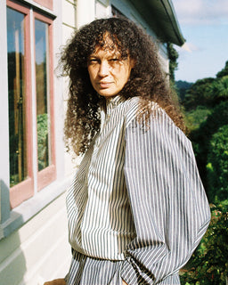 A woman with long, curly dark hair stands outdoors in front of a house, wearing a loose, high-neck striped top and matching bottoms. The outfit features vertical stripes in gray and off-white.