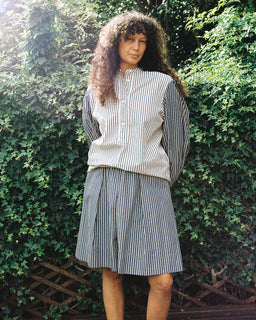 A woman with long curly hair stands outdoors in front of a leafy green hedge, wearing a matching striped button-up shirt and pleated skirt set.