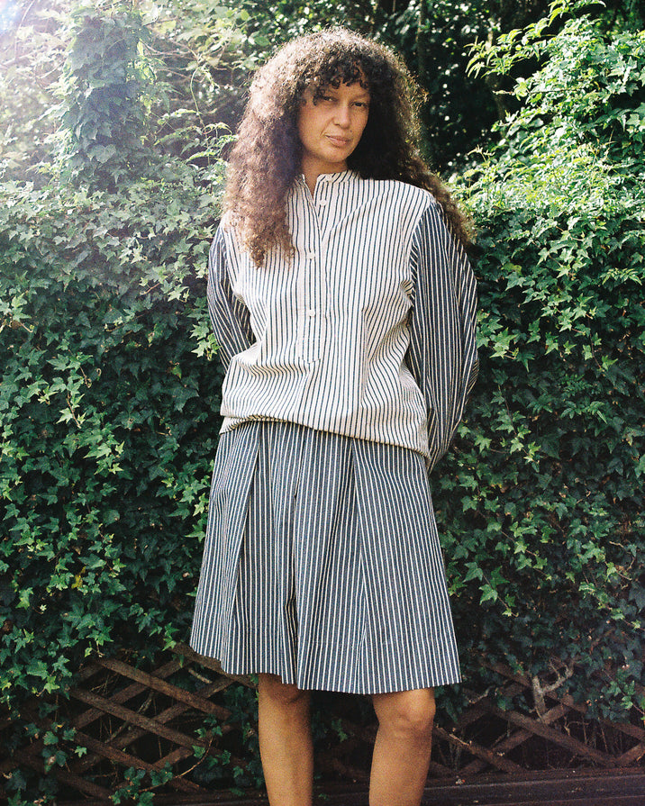 A woman with long curly hair stands outdoors in front of a leafy green hedge, wearing a matching striped button-up shirt and pleated skirt set.